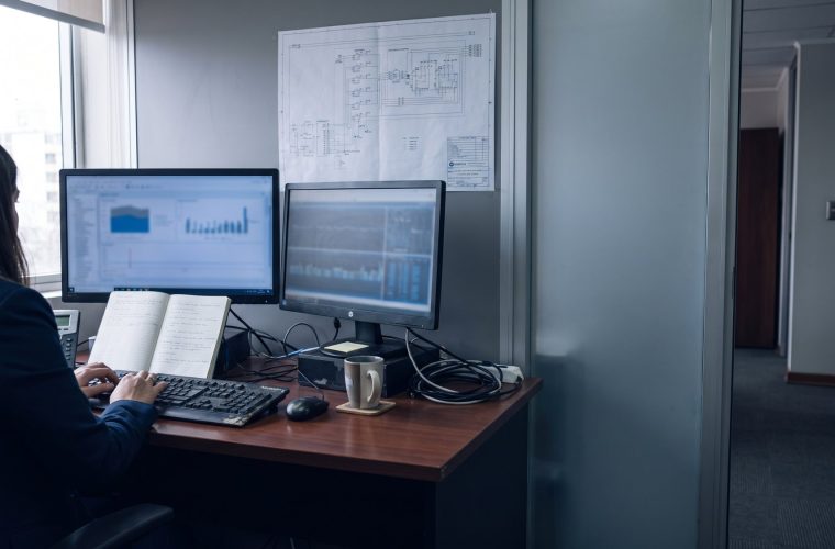 Puesto de trabajo de tecnologia con dos monitores, un cuaderno con anotaciones y las manos de una persona trabajando